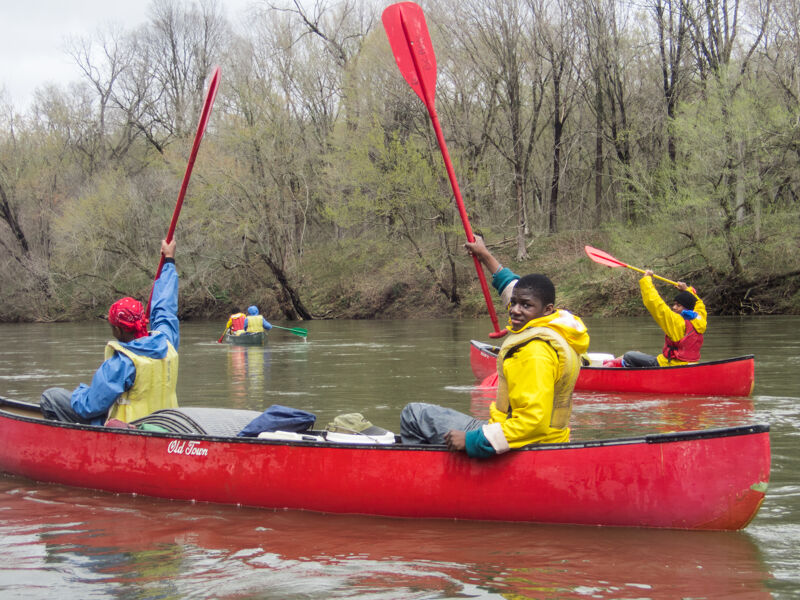 The image shows three red canoes on a river. Each canoe has one or two people in it, all wearing life jackets. They are holding red paddles in the air. The river is surrounded by trees with bare branches, suggesting it might be early spring or late fall. The water is calm and reflects the colors of the sky and trees.
